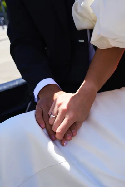 Couple holding hands, engagement ring close-up catching light as their fingers interlace on sunlit outdoor pavement.