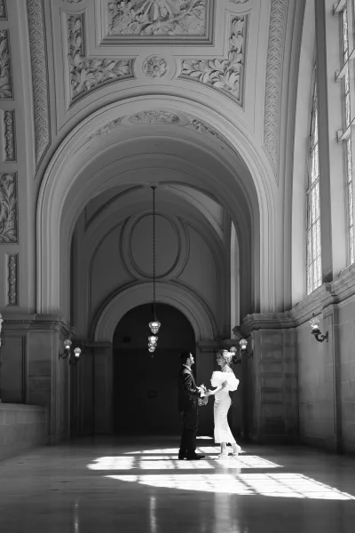 Couple portrait of a bride and groom holding hands, the bride clutching a small bouquet in an arched hallway with an ornate ceiling.