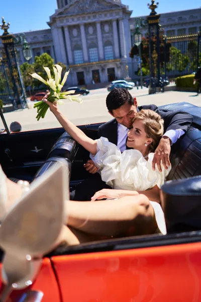 Getaway car moment captured as a just married car photo: newlyweds cuddle, bride raising a calla lily bouquet by a stone facade.