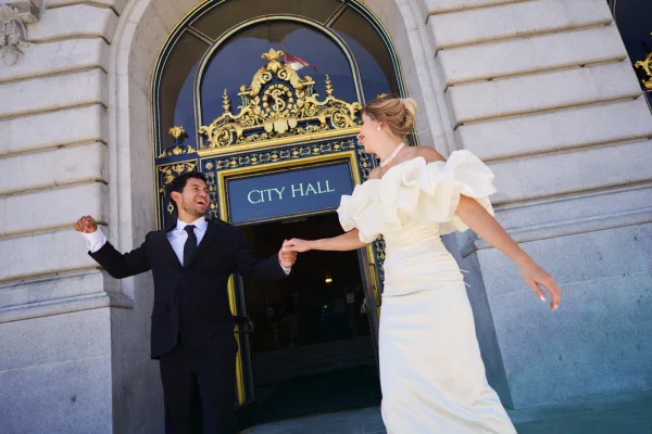 City hall elopement couple sharing a joyful wedding exit beneath an ornate stone archway, bride in ruffled sleeves leading her partner.