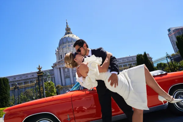 Wedding kiss portrait of a bride and groom dip beside a classic convertible, framed by a historic dome and wrought iron gate under blue sky.