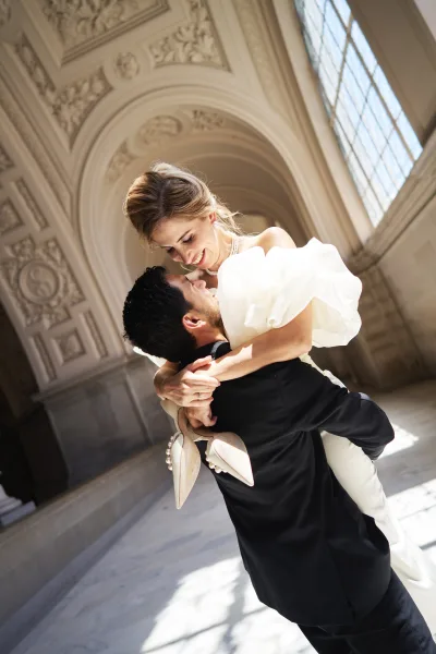 Couple portrait with the bride lifted by the groom, her pearl necklace gleaming, framed by ornate arched windows over a polished marble floor.