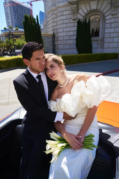 Couple portrait of a bride and groom embrace beside a historic building facade, the bride holding a calla lily bouquet.