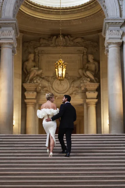Couple portrait of a bride and groom walking up a grand staircase beneath an ornate ceiling and chandelier, her off-the-shoulder gown.