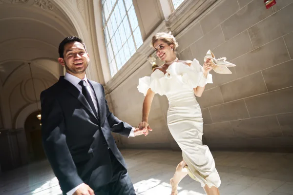 Couple portrait of a bride and groom running hand in hand through an arched hallway with large windows, her pearl necklace catching soft light