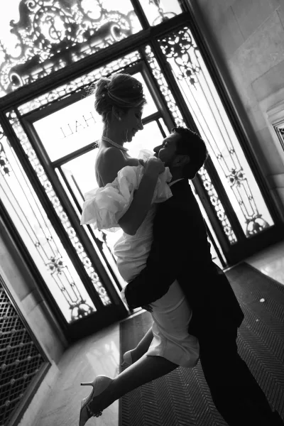 Groom lifting bride in a couple portrait, her ruffled sleeves flaring as they smile before ornate glass doors with wrought iron.