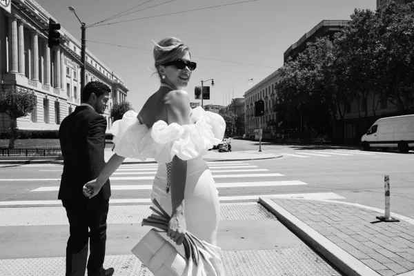 Couple portrait of a bride and groom walking hand in hand across a city crosswalk, the bride in sunglasses, buildings and trees lining the street