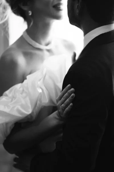 Black and white couple portrait in a romantic wedding embrace, the bride’s engagement ring catching light as they lean close beside a window.