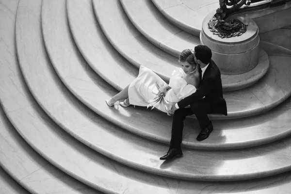 Couple portrait of bride and groom sitting on curved stone steps, her minimalist bouquet resting in her lap beneath an ornate railing.