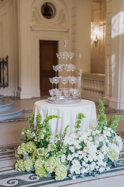 Champagne tower as a wedding champagne display with florals at the base, on mosaic tile amid white walls and classical architecture.