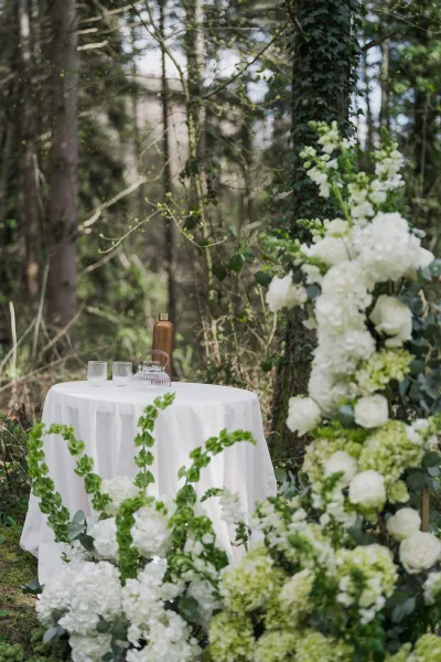 Ceremony table styled for outdoor ceremony decor, adorned with white florals, set against mossy forest trees and ivy for a serene woodland feel.