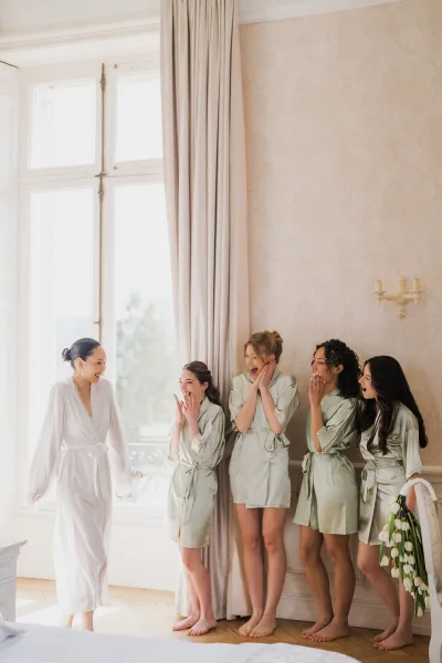 Bride and bridesmaids during getting ready photos laugh together in matching satin robes by a large window with curtains and wallpaper.