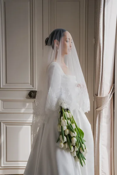 Bridal portrait of a bride under veil, holding a white tulip bouquet, in a minimalist long-sleeve gown with classic, elegant poise.