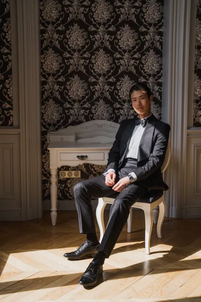 Groom portrait showcasing tuxedo groom style as he sits, adjusting his bow tie against damask wallpaper and wall paneling.