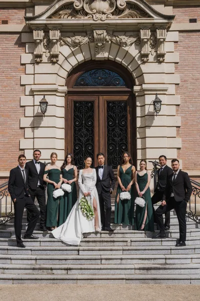 Wedding party portrait of the bridal party on steps, emerald green bridesmaid dresses framed by a brick facade and arched wrought iron doors.
