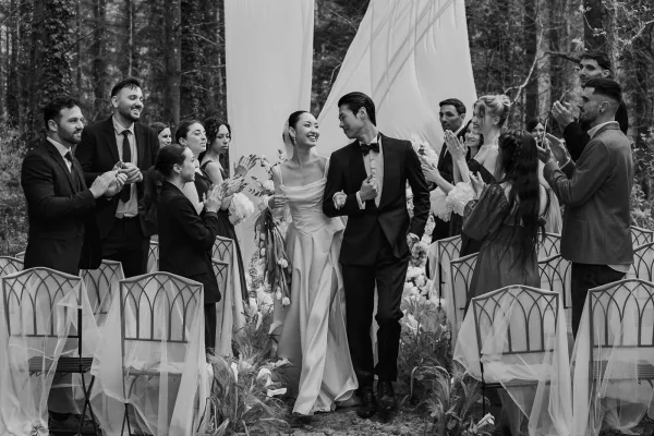 Wedding recessional with wedding guests clapping as arm-in-arm newlyweds walk past aisle flowers under forest trees, grinning at each other.
