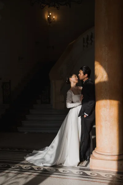 Couple portrait of a bride and groom embrace on a grand staircase with wrought iron railing, her gown featuring a square neckline.
