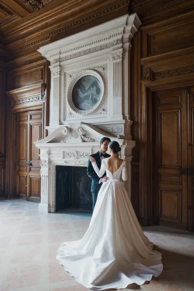 Couple portrait of a bride and groom embrace by an ornate fireplace, her satin ballgown with a long train framed by carved wood paneling.