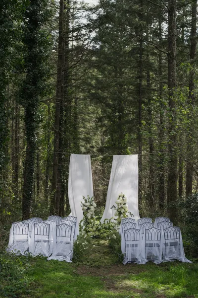 Outdoor ceremony setup for a woodland wedding ceremony with white draping, framed by forest trees over a grass lawn.