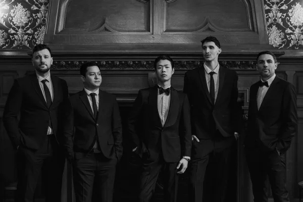Groomsmen portrait of the groom with groomsmen in classic tuxedos, posed by a wood-paneled fireplace mantel and floral wallpaper.