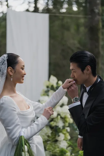 Couple portrait as the groom kisses the bride's hand, her lace veil trailing softly against forest trees and a white fabric backdrop.