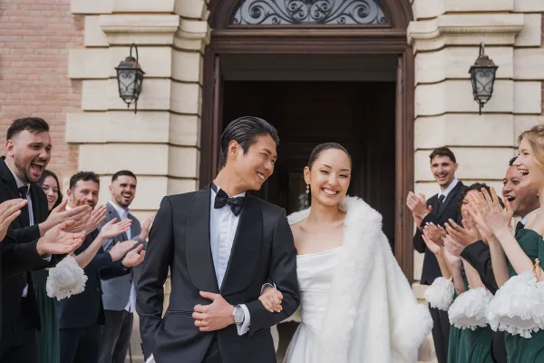 Wedding recessional as the bride and groom exit arm in arm, her white faux fur wrap catching the light, past a stone facade and arched doorway.