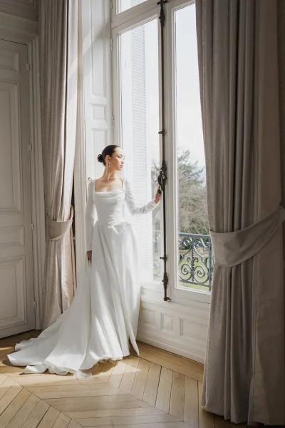 Bridal portrait of a bride by window in a square-neck, long-sleeve wedding gown, framed by tall curtains and a balcony with trees beyond.