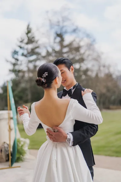 Couple portrait in formal wedding attire, bride with a sleek low bun and delicate hair pins on a stone patio by trees.