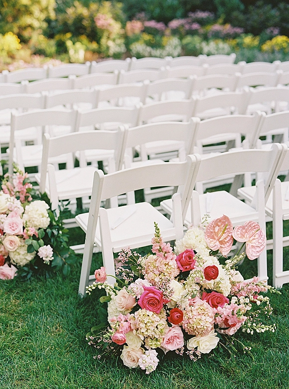 Ceremony seating with white folding chairs lining a lawn aisle, with low rose, hydrangea, and greenery clusters against garden foliage