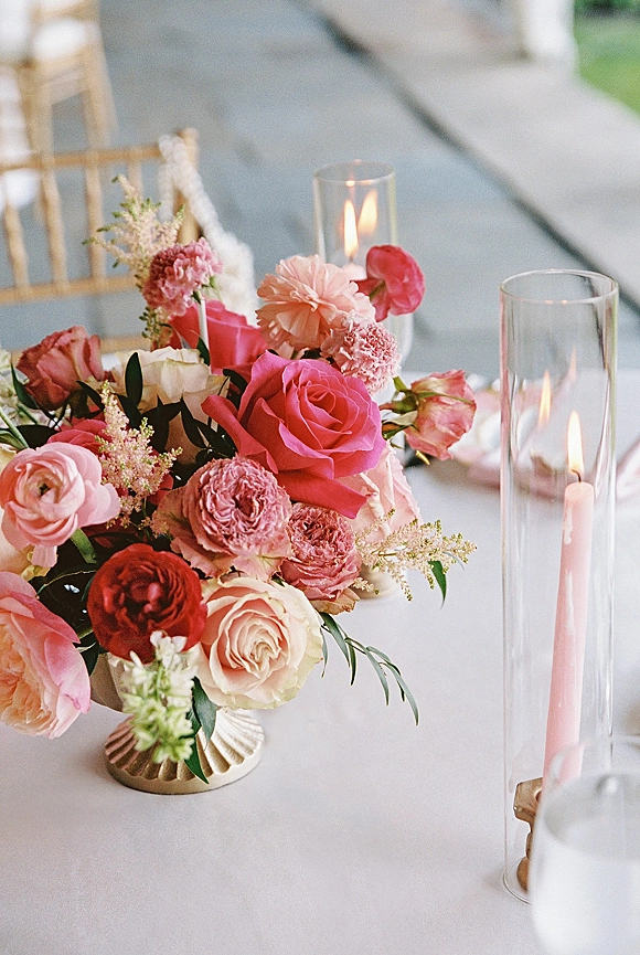 Reception centerpiece with a wedding floral centerpiece of pink roses and mixed blooms in a pedestal vase, flanked by taper and glass hurricane candles on an outdoor lawn table