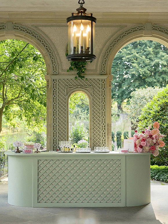 Wedding bar setup with champagne coupes and cocktail glasses on a pastel green counter, framed by stone arches and a hanging lantern accent
