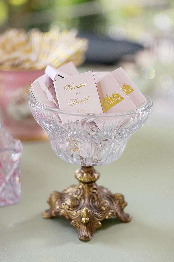 Wedding favor display featuring personalized matchboxes wedding favors in a glass pedestal bowl with gold foil printing on a tabletop, blurred decor behind