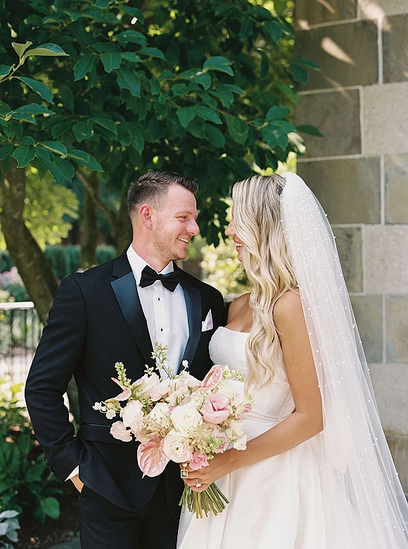 Couple portrait of bride and groom smiling at each other, bride holding a pink and white bouquet, with veil, tuxedo, stone wall garden backdrop