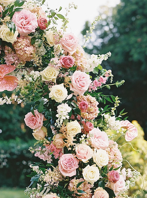 Wedding ceremony florals on a floral arch arrangement with pink and ivory roses, peonies, hydrangea, and greenery against garden trees