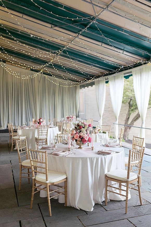 Reception tablescape with round wedding table setup, blush napkins, gold Chiavari chairs, floral centerpieces and taper candles under patio string lights