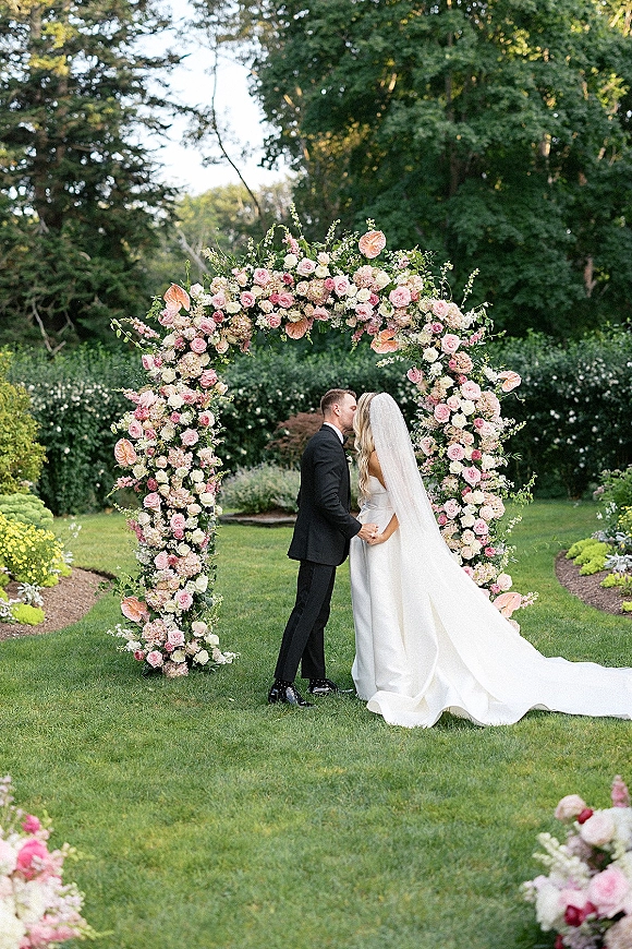 Wedding kiss portrait of bride and groom kissing under arch with pink-white roses, hydrangeas and greenery on a garden lawn