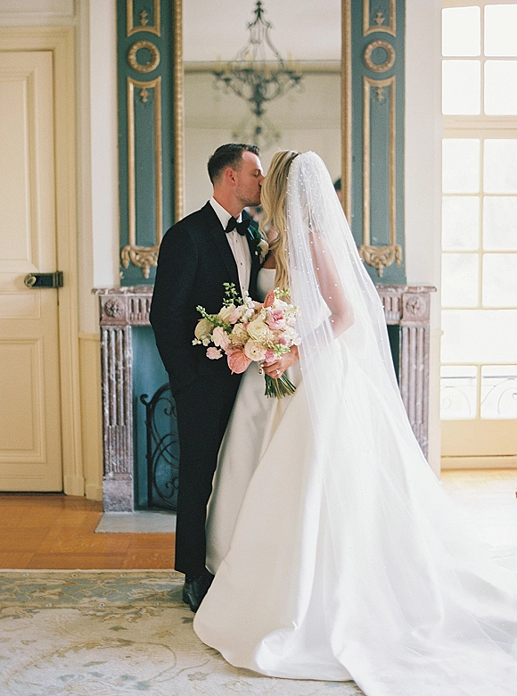 Wedding kiss as bride in strapless gown and long veil kisses tuxedoed groom, bouquet in hand, beneath chandelier by ornate mirror fireplace