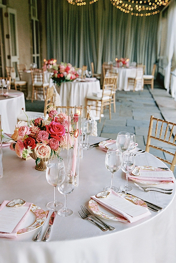 Reception tablescape with round wedding table setup, rose floral centerpieces, taper candles in glass vases under string lights on a covered terrace