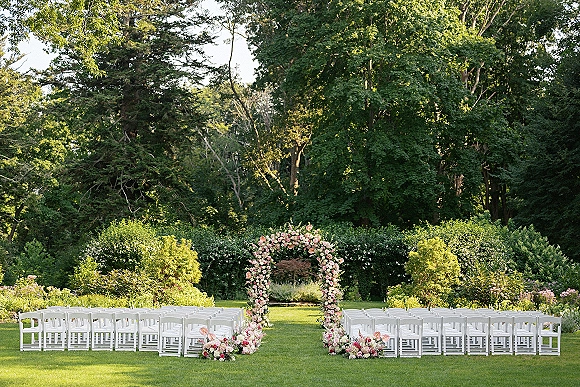 Ceremony setup for outdoor wedding ceremony with a circular floral arch of pink roses and white flowers, framed by white chairs on a sunny lawn