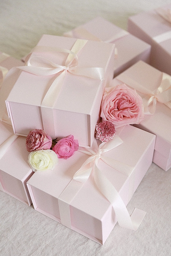 Wedding favor boxes in pink arranged on linen with satin ribbon bows and rose, ranunculus, and carnation accents in a flat lay