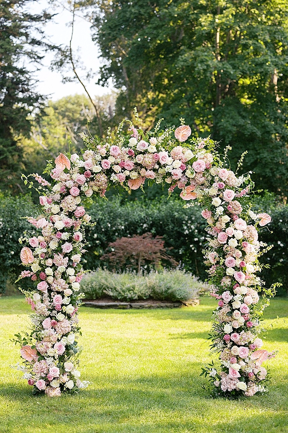 Wedding floral arch with roses, hydrangeas, greenery, and anthurium arranged on a round outdoor ceremony arch on a garden lawn