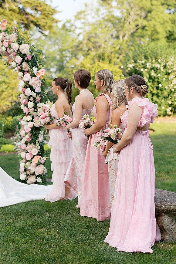 Bridesmaids portrait in pink bridesmaid dresses holding bouquets beside a floral ceremony arch on a garden lawn with hedges and a stone bench