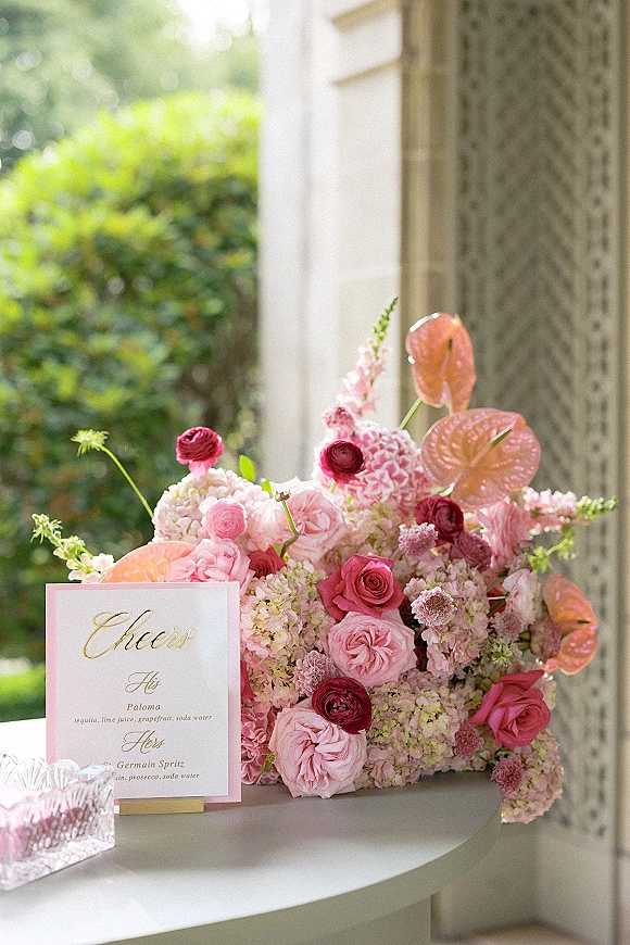 Cocktail bar decor with a wedding signature drink sign in an acrylic holder beside a pink floral arrangement on a glass tray outdoors