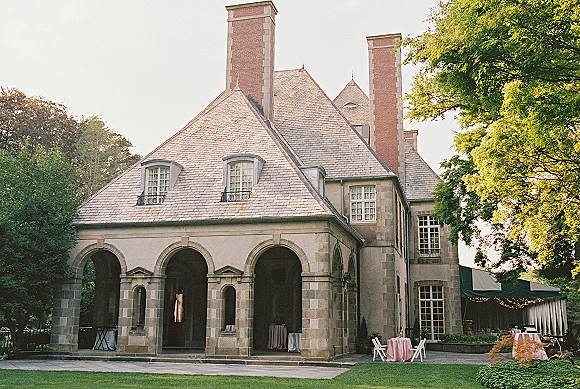Wedding venue exterior with round cocktail tables under bistro string lights beside a stone manor house and arched patio on a green lawn