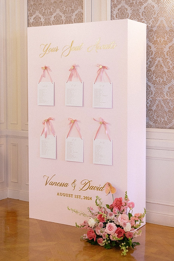 Wedding seating chart with seating chart display cards in gold calligraphy, pink ribbon escort cards, and lush florals against patterned wallpaper