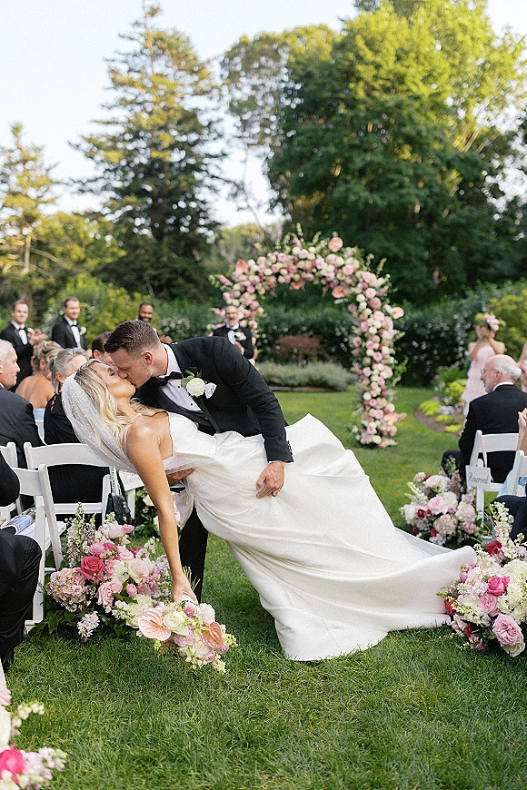 Wedding kiss moment as the groom dips the bride for a ceremony kiss dip beneath a pink and white floral arch on a garden lawn aisle