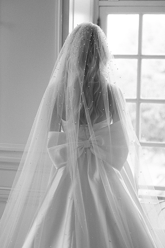 Bridal portrait of a back view bride in a strapless satin ball gown with a pearl wedding veil and bow sash, lit by soft window panes indoors