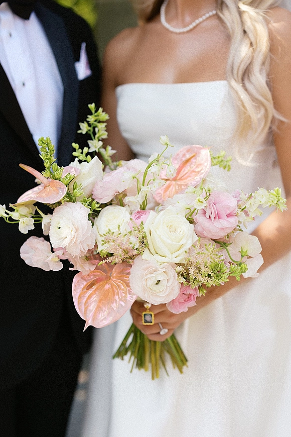 Bridal bouquet of pink and white blooms with anthurium, roses, ranunculus, and greenery held by bride in strapless dress outdoors