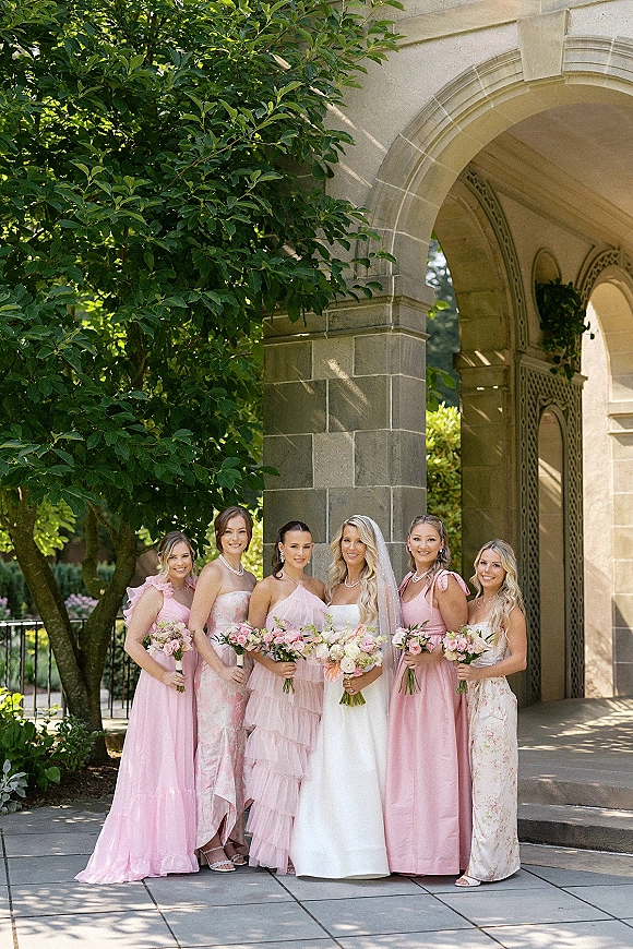 Bride with bridesmaids in pink dresses holding pastel bouquets, the bride in a strapless gown and veil beneath a stone archway