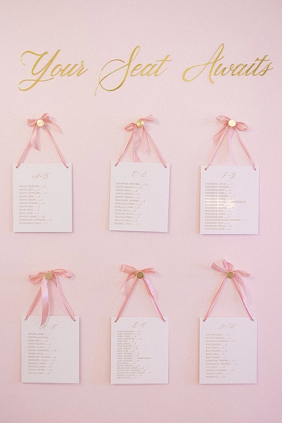 Wedding seating chart with gold calligraphy lettering on cards, each hung with pink ribbon bows and round fasteners against a blush wall backdrop